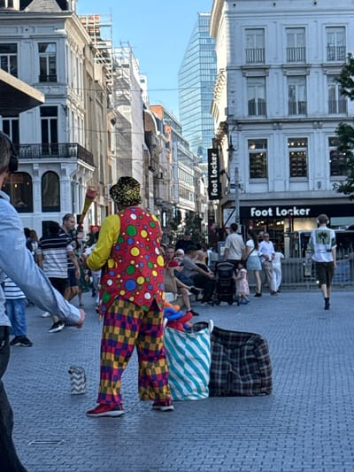 The image captures a street scene in Brussels, Belgium, specifically on Rue Neuve. A street performer dressed as a clown, wearing a colorful polka-dot vest and checkered pants, is the central figure. The clown is holding a juggling prop. Several other individuals are visible in the background, some walking, others sitting. A Foot Locker store is identifiable by its signage. The environment includes traditional European architecture on the left and a modern glass skyscraper in the distance on the right. The ground is paved with cobblestones. The scene appears to be set during daylight, with a clear blue sky.
