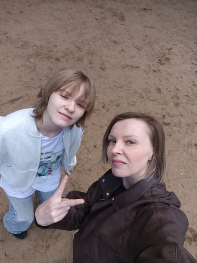 Two individuals are visible in this high-angle shot, likely a selfie. One is a younger person with light brown hair, bangs, and wearing a white zip-up jacket over a t-shirt with an indistinguishable graphic. They are making a peace sign with their hand. The second person, presumably the photographer, is an adult with shorter blonde hair and a dark brown leather jacket. The background consists of a dirt-covered area, possibly a sandy beach or unpaved ground. The location is alleged to be the city of Chaykovsky, Russia. No other objects or interactions are visible.