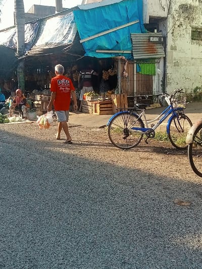 The scene takes place in Pati, Indonesia, at the coordinates -6.756 latitude and 111.038 longitude. A man wearing an orange shirt with the words "PITI" on the back, and blue shorts, walks away from the camera, carrying a plastic bag with produce. The man is walking on a paved road in front of a small shop with a blue tarp awning. Behind the shop is a white wall with "BELAKANG 6" written in black. Two women sit on the ground near the entrance to the shop. One woman is wearing a blue shirt, the other is wearing a white head scarf. A blue and white bicycle is parked on the side of the road in front of the shop. A second bicycle is partially visible on the right side of the image. The shop appears to be a fruit stand, with boxes of produce in front. The pavement is covered with small rocks.  The sun is shining brightly. The day is clear and the sky is blue.  The scene is a typical street scene in Pati, Indonesia.