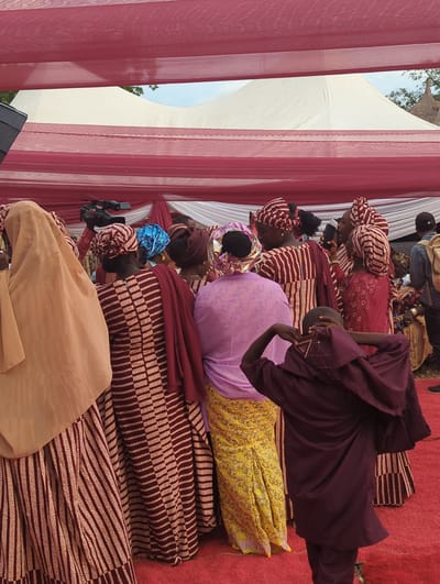 This image depicts an outdoor gathering taking place in Kabala, Nigeria, under a temporary canopy structure. The ground is covered with a red carpet.
The scene is populated by approximately 10-15 visible individuals, primarily women, dressed in various forms of traditional attire. Many women wear patterned dresses and matching head wraps in shades of maroon, red, and light tan, featuring vertical stripes or geometric patterns.
- On the far left, a woman is seen from the back, wearing a light tan head covering and a vertically striped dress.
- In the center-left, a woman is visible from the back, wearing a maroon and tan patterned dress with a dark red shawl draped over her shoulders. Her head is covered with a matching patterned wrap.
- Slightly behind her, a person is operating a black JVC video camera on a monopod or tripod, pointing towards the right.
- In the central foreground, another woman is viewed from the back, wearing a light purple top and a yellow patterned skirt, with a matching purple head wrap.
- To the right, several other women in similar attire are present, some engaged in conversation. One woman in a red dress is holding a black smartphone to her ear. Another woman wears a distinctive bright blue head wrap.
- In the immediate foreground on the right, a young individual, possibly a child or adolescent, is observed from behind, wearing a dark maroon tunic with light embroidery near the neckline and matching pants. Their hands are raised to their head, seemingly adjusting headwear.
- In the far right background, a person with a black baseball cap and a light-colored backpack is partially visible.
The overhead canopy consists of alternating red and white fabric sections. In the background, a large white marquee-style tent is visible, with parts of a blue sky and green foliage, including the top of a traditional straw-roofed structure, peeking out from behind it.