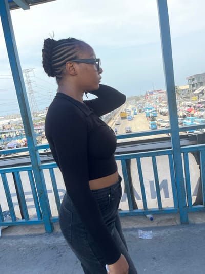 The image depicts a female individual standing on a pedestrian bridge overlooking a busy urban street in Lekki, Nigeria. The subject, visible from mid-thigh up and facing right in profile, has dark skin, hair styled in cornrows pulled back into an updo, and wears dark rectangular sunglasses. She is dressed in a black long-sleeved, collared, cropped top with front button details and dark-wash jeans. Her left arm is raised with the hand positioned behind her head/neck.

The bridge features blue metal railings with vertical bars. On the concrete bridge floor near the bottom right, a clear plastic water bottle and a crumpled plastic bag are present.

Below the bridge, a multi-lane expressway is visible with moderate to heavy vehicle traffic, including passenger cars, a prominent yellow truck, and numerous light blue public transport buses. To the right of the expressway, a dense market area is established, characterized by a multitude of white and brightly colored umbrellas and canopies covering numerous stalls, with multi-story buildings beyond. To the left of the expressway, scattered buildings and a tall electricity transmission tower (pylon) are visible. The sky is light grey-blue, indicative of an overcast or hazy atmospheric condition. The individual appears to be observing the activity below the bridge.