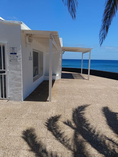 The image depicts a bright and sunny coastal scene in Puerto del Carmen, Spain, featuring a pristine white building alongside a paved outdoor area overlooking the ocean. The single-story building, likely a public facility or information center, has a white gate on the left and a large rectangular window.
Extending from the building are two white pergolas; one is attached to the building, and another, smaller one stands further out, casting distinct striped shadows on the light brown, pebble-dashed paving. Large, intricate shadows of palm tree fronds are prominently cast across the foreground and the paved surface, indicating strong, direct sunlight.
Beyond the paved area, a low black stone wall separates the land from the vast, deep blue expanse of the Atlantic Ocean. The horizon is clear, and the sky above is a brilliant, cloudless blue. Visible palm fronds, dark green against the bright sky, frame the upper right portion of the image.
Two signs are affixed to the white building. On the left, a blue and white plaque displays "Calidad Turística / Tourist Quality 2024" and lists opening hours as "Lunes a Domingo de 10 a 18 horas" (Monday to Sunday 10 am to 6 pm). Next to the window, another sign reads "AYUNTAMIENTO DE TÍAS / CONCEJALÍA DE TURISMO" (Municipality of Tías / Department of Tourism), suggesting the building's function as a tourist office.
The scene conveys a peaceful and pleasant atmosphere, characteristic of a sunny day at a coastal resort. The sharp shadows indicate it is midday or early afternoon.
