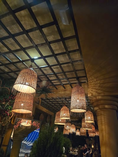 The image captures the interior of a dimly lit restaurant with a rustic ambiance, possibly located in Sofiivska Borshchahivka, Ukraine. The ceiling features a dark wooden lattice structure with cream-colored panels, from which numerous woven basket-like lamps hang at varying heights, casting warm, intricate shadows on the walls and ceiling. In the foreground, to the left, an archway reveals a reflection of bright string lights against a dark blue background, suggesting an outdoor or decorative element. Above the arch, a sign with Cyrillic lettering is partially visible. To the right, a large, fluted column stands prominently. In the background, several people are gathered around tables, engaged in conversation. Lush green foliage and decorative elements contribute to the overall atmosphere.