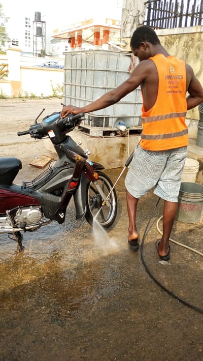 In Adankolo, Nigeria, an outdoor scene depicts a male individual pressure washing a dark red Haofeng 125 underbone motorcycle. The man, with dark skin, wears an orange sleeveless reflective vest inscribed with "BWLYRICS" and patterned light-colored shorts, along with black flip-flops. He holds a black and silver pressure washer, directing a stream of water at the motorcycle's front wheel. The motorcycle is supported by its kickstand on a wet, paved surface. Directly behind the man stands a large, grey, framed industrial water tank. To his right, two plastic buckets, one white and one grey, are visible. The background features a low white wall, a multi-story building, a distant water tower, and a structure with orange architectural details, suggesting a commercial building.
