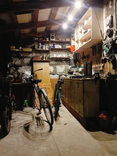 The image depicts the interior of a cluttered workshop or garage, likely located in Slutsk, Belarus. The space is dimly lit, with artificial lights casting shadows across the concrete floor and the various items stored within. In the foreground, two bicycles are parked, their wheels casting long shadows. One bicycle, to the left, is partially obscured by another object. To the right, a second bicycle stands upright.
The workshop is equipped with shelving units filled with containers, tools, and other assorted items. A workbench or counter runs along the far wall, laden with equipment, including what appears to be a power tool and a battery pack. The walls are rough, likely made of concrete or stone, and show signs of age. Wooden beams are visible on the ceiling, supporting what appears to be some sort of insulation or paneling. Various tools are hung on the wall behind the workbench, suggesting a space used for repairs or crafting. The overall impression is one of a functional, but somewhat disorganized, workspace. There are no people visible in the image.