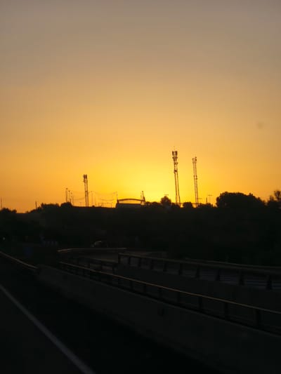The image depicts a sunset scene with a vibrant orange sky over Paterna, Spain. In the foreground, a dark silhouette of a highway overpass with guardrails runs horizontally across the lower part of the frame. The highway lanes are visible on the left side. Beyond the overpass, a dark, undulating landscape of trees and hills is silhouetted against the bright sky. Several tall, slender structures, possibly telecommunication towers or lighting poles, rise from the hilltops. A domed structure, possibly part of a sports facility, is also visible on the horizon. The overall impression is one of a transition from day to night in an urban or semi-urban environment.