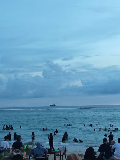 The image shows a beach scene at Monikeun, Indonesia. The sky is overcast with blue-gray clouds. In the distance, a cargo ship is visible on the horizon. Numerous people are in the water, some standing, others swimming. In the foreground, people are sitting at tables, with fresh coconuts on the table. One table has the number "31" visible. Some individuals are taking photos.