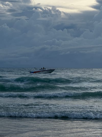 The image depicts a vertical, split-screen view of a cloudy sky on the left and a choppy ocean on the right, bisected by a sailboat. The sky on the left is filled with dramatic, dark, and heavy clouds, suggesting a stormy or overcast day. Light filters through in some areas, creating a dramatic contrast. The right side of the image shows the ocean with active, breaking waves, indicating a rough sea.

A small, white sailboat with red and blue accents is positioned in the middle, partially on the water and partially against the dark clouds. Two figures are visible on the boat, suggesting they are sailing or operating the vessel. The boat appears to have text on its side, which is partially obscured and difficult to read clearly due to the image resolution and angle. However, some Thai characters are discernible.

The scene conveys a sense of movement and potentially challenging conditions due to the rough seas and dark clouds. The lighting suggests it could be late afternoon or early morning, with the sun low on the horizon, contributing to the dramatic sky. The location context of Karon, Thailand, hints at a tropical coastal environment. The overall mood is one of nature's power and perhaps human interaction with it.