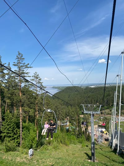 A slightly tilted, eye-level shot of a ski lift in the Прикамский area of Russia. A person, wearing a dark-colored shirt and pants, sits beside a child wearing a pink-colored shirt and beige pants in the ski lift chair. Both figures appear to be looking to the side. The chair they are sitting in has a gray frame. In the background, a dense forest and sky are visible. Some buildings are visible. Above the lift chair, the sky is blue with clouds. In the foreground, a thick black cable of the ski lift is visible. Other cables stretch across the sky in the background.