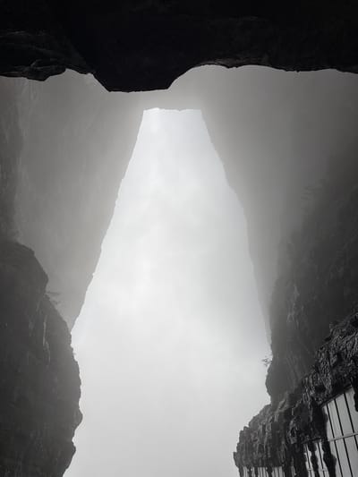 The image depicts a view upwards through a gap in rock formations. Dense fog obscures much of the landscape, but the silhouettes of mountains can be discerned in the middle ground. In the lower right and left corners are dark, rocky cliffs. The upper right corner shows a dark, geometric structure, possibly a building or constructed platform, atop a rock face. The photograph was taken in Yongding District, China.