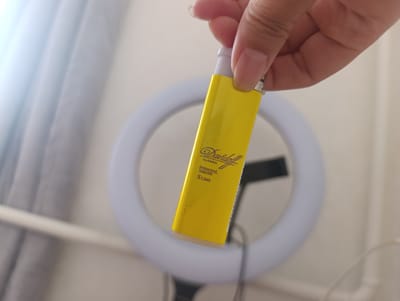 A person's hand is holding a yellow lighter. The lighter has the brand name "Davidoff" written in script, with "CLASSIC", "Switzerland SINCE 1970", and "SLIMS" written below it. The background of the image is blurred but shows a ring light and what appears to be a curtain. The lighting suggests it might be daytime. The scene is indoors, possibly in a home or studio setting. No specific emotions are conveyed.