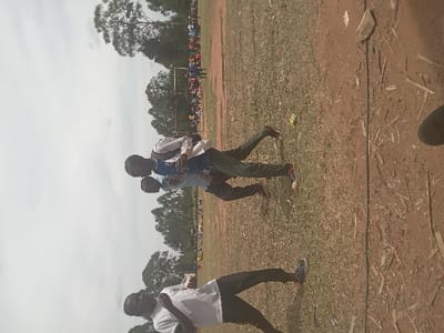 The image shows three men standing on a grassy field in Lwandeti, Kenya. The location is identified by its latitude (0.545) and longitude (34.834). The men are all wearing casual clothes and are looking at the camera. Two of the men are facing the camera, and one is standing behind and to the left of the other two. The man standing behind and to the left is holding a white bag, and his right hand is on the shoulder of the man in front of him. Behind the men, a group of people are in the distance, possibly watching a game. There is a wire or cable running diagonally across the field. The scene is a mix of grass and dirt. The sky is cloudy.
