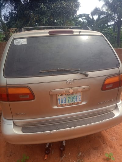 The image displays the rear exterior of a champagne-colored Toyota Sienna XLE minivan, positioned on a reddish-brown dirt ground in Alor, Nigeria. The vehicle features a dark-tinted rear window with a single horizontal wiper blade. A white sticker on the upper left corner of the rear window reads "GREAT IS JEHOVAH" and "GOD'S KINGDOM GOVERNS." A Nigerian license plate, registered to "ABUJA CENTRE OF UNITY" and bearing the alphanumeric sequence "YAB-289EE," is mounted centrally on the tailgate, beneath the Toyota emblem. The rear bumper exhibits scuff marks and paint chipping, with a black rubber protective strip across its width. Two dark-colored glass bottles are visible lying on the ground directly behind the bumper, partially tucked underneath the vehicle. The background consists of dense tropical vegetation, including large-leafed trees and palm trees, beneath an overcast sky. No individuals are present in the scene.