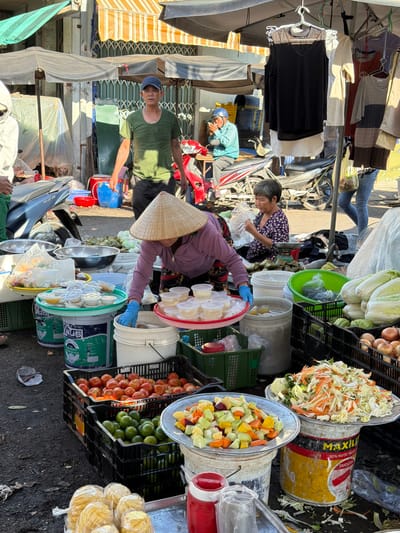 This image captures a bustling street market scene in Nha Trang, Vietnam. A man wearing a green t-shirt and a blue baseball cap stands in the center, appearing to be a vendor. To his right, a woman in a light blue shirt and helmet is seated on a motorcycle, smoking. In the foreground, a woman with a conical hat is arranging small containers of food. Several stalls are visible, displaying a variety of produce including tomatoes, limes, and what appear to be cabbages and onions. Clothing and other goods are also displayed for sale. The overall atmosphere suggests a busy commercial area with daily life unfolding.