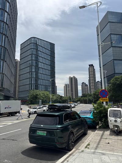 In the Haizhu District of China, the image depicts a street scene with contemporary architecture. On the left, a large, white box truck with Asian characters on its side is parked. Two individuals, one wearing a white shirt and pants and the other a black shirt and pants, are walking across the street, with the person in white appearing to be looking towards the left. A dark green SUV is parked in the foreground, facing away from the camera, with a license plate that reads "A•F37899". The SUV has a roof box. To the right of the SUV, a turquoise car is parked, followed by a white three-wheeled vehicle that resembles a tuk-tuk. Tall, modern glass-fronted buildings dominate the background, with a light-colored sky overhead. A street lamp with two lights is visible on the right side of the frame. A circular red and blue sign with a diagonal cross indicates a prohibition. The road has white lane markings. Green trees are present along the street and sidewalks.