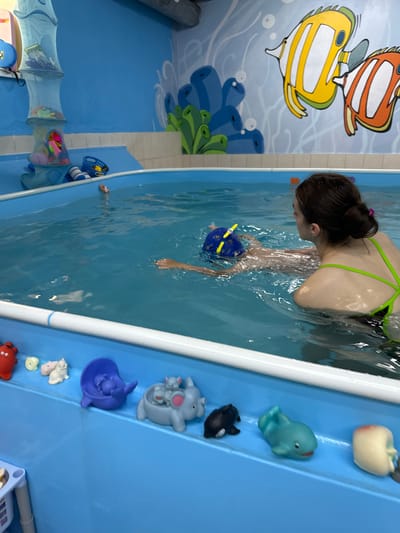 This media file shows a young woman swimming in a bright blue pool. She is wearing a green bikini and has her dark hair tied up in a bun. She is holding a blue and yellow swim toy. The pool is surrounded by blue walls decorated with colorful cartoon fish and coral. On a blue slide next to the pool are several rubber toys, including a white apple, a blue dolphin, a gray hippo, and a black and white seal. There are also some mesh toy bags hanging on the wall. The scene appears to be indoors, possibly at a swimming class or a pool facility. The lighting is bright, suggesting daytime. The overall impression is one of a fun and playful environment.