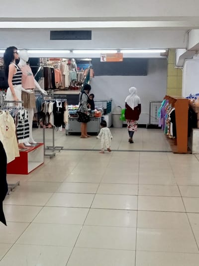 The image shows a shopping mall interior in Pati, Indonesia, located at coordinates -6.757, 111.039. There are four people, two women and two children, visible in the scene. One woman is wearing a hijab, while the other is wearing a black top and jeans. One of the children is wearing a white dress, and the other is wearing a white dress and a black and white striped top. The women are walking towards the right side of the image, while the children are standing still. One woman is carrying a baby in her arms. The scene includes numerous clothing racks filled with clothes, a cash register, and a display of clothing. There is also a red and white shelf in the foreground. The floor is tiled in white and beige, and the walls are painted white. The ceiling is white with visible exposed support beams.
