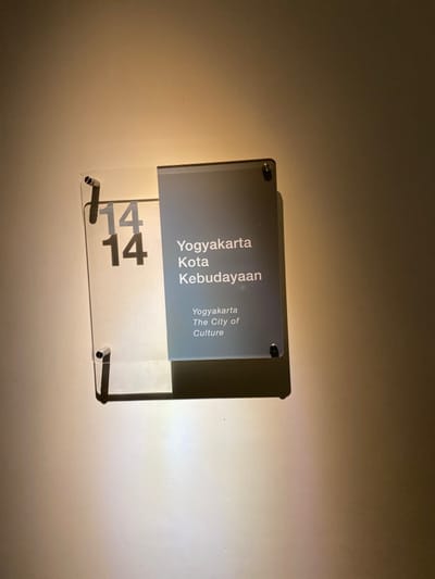 The image shows a sign mounted on a wall. The sign is made of clear acrylic and is a rectangular shape. The sign has a gray rectangle in the center that has black text that reads "Yogyakarta Kota Kebudayaan" and "Yogyakarta The City of Culture." The number "14" is displayed on the left side of the sign, in large black font. The sign is mounted on a light-colored wall. The image was taken in Banguntapan, Indonesia, which is located at latitude -7.8 and longitude 110.407. The image was likely taken indoors.