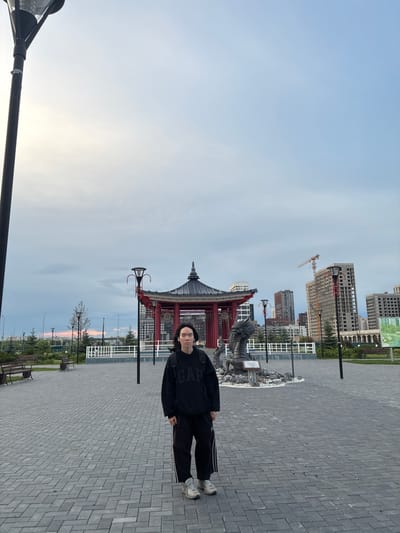A person stands in a paved outdoor area with a large open space and buildings in the background. The person is wearing a black hoodie with white lettering and black pants. They are looking towards the camera. The scene appears to be a public space with benches and lampposts visible. In the mid-ground, there is a red and grey structure resembling a pagoda. Construction cranes and unfinished buildings suggest an ongoing urban development in the background. The sky is a pale blue with some clouds, indicating it is likely daytime, possibly late afternoon or early evening. The lighting is soft. There is text on the hoodie that appears to read "GAP". The setting seems to be a modern urban environment in Tyumen, Russia, with elements of cultural architecture integrated into the cityscape.
