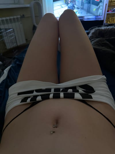 This is a close-up, vertically oriented photograph featuring a person's midriff and upper thighs. The individual is wearing white shorts with a black striped logo and black underwear that is partially visible. The person has a belly button piercing. The lighting is dim, suggesting an indoor setting, possibly at night. The background is blurred, but hints of objects like a gaming machine or arcade cabinet on the left and what appears to be a radiator or shelving unit on the bottom right can be discerned. The overall tone of the image is intimate and personal, focusing on the details of the body and clothing.