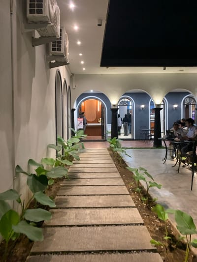The image shows a walkway lined with large, green, leafy plants, leading to an entrance of a restaurant or cafe with arches. The entrance leads to a seating area with a few people seated at tables. The walkway is made of stone and leads to a courtyard. There are two large, white air conditioners on the wall of the building, which is painted white. The image was taken at a location in Malang, Indonesia with latitude -7.955 and longitude 112.634.