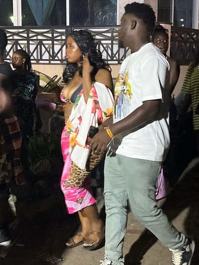 A group of people is present in the scene. A woman wearing a white, floral dress with a leopard print panel stands next to a man in a white t-shirt and green pants. He is holding a phone. Another woman is partially visible in the background, wearing glasses. The setting appears to be outdoors, possibly at an event or gathering.