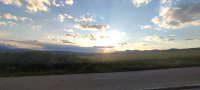 The image shows a rural landscape at sunset. The sky is a vibrant blue with scattered white and gray clouds, illuminated by the setting sun which is partially obscured by clouds, casting rays of light across the scene. In the foreground, there is a paved road with a white line marking its center. Beyond the road, a line of dark green bushes and trees stretches horizontally across the mid-ground, separating the road from a large, open field of green grass. In the distance, rolling hills or mountains form a silhouette against the horizon, their dark shapes contrasting with the bright sky. The overall impression is one of natural beauty and tranquility. The location is Momin Prohod, Bulgaria.