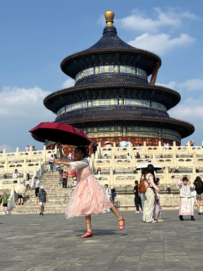 An outdoor, eye-level, medium-wide shot captures the Hall of Prayer for Good Harvests in the Temple of Heaven, Dongcheng District, China, under a clear blue sky with scattered white clouds. A young girl in a pink, layered dress and red shoes is the prominent subject in the foreground, holding a partially opened red umbrella above her head. Her left leg is extended upwards and forward, her right leg bent, and her body is turned to her right, suggesting a dance-like pose.
Behind her, a large multi-tiered, ornate temple structure dominates the background, its dark blue, multi-layered roof adorned with gold finials. White stone balustrades and a long flight of stairs lead up to the temple, with numerous people scattered throughout the scene. Some individuals are ascending the stairs, while others are standing on the platforms, many holding umbrellas to shield themselves from the sun. The ground in the foreground is paved with large, grey stone tiles. The lighting suggests it is a sunny day.