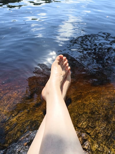 A pair of bare feet, belonging to an individual with fair skin, are submerged in dark, calm water. The feet are positioned with the left foot crossing over the right, and the toes are visible. The water is clear enough to see the rocky, golden-brown bottom beneath, with some darker shadows indicating depth and submerged rocks. Sunlight reflects off the water's surface, creating shimmering patterns. The scene appears to be a natural setting, possibly a lake or river bank. While Norrköping, Sweden, is mentioned as a potential location, the image itself does not contain any specific visual cues to confirm this.