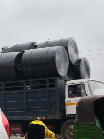 In Aba, Nigeria, the image captures a flatbed truck carrying large, black cylindrical tanks secured with green straps. The truck has a black bed and a white cab with a yellow stripe. A partially visible green auto-rickshaw is to the right of the truck. To the left of the truck is a white car with a red taillight. A person wearing a yellow shirt and black pants is crouching near the rear of the truck. Overcast sky.