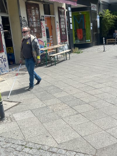 In Berlin, Germany, a man with a white beard and sunglasses walks on a sidewalk holding a white cane. He is wearing a grey and patterned jacket over a dark t-shirt with a yellow smiley face logo. He has on blue jeans and dark shoes. The sidewalk is paved with large grey tiles. Behind the man, there is a building with graffiti on its facade. Several wooden picnic tables are set up in front of the building. Further down the street, there is a shop with a green facade and a brightly colored display visible through the window. A few trees are planted along the street. The scene appears to be a public street with shops and outdoor seating.