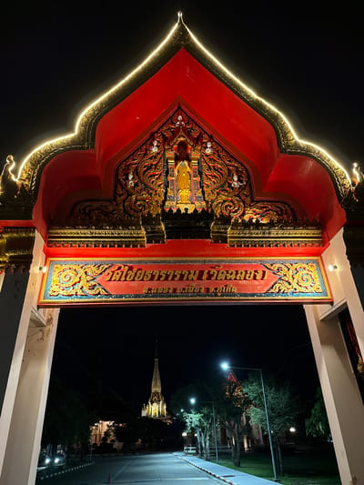 An image taken at night depicts the entrance to a Buddhist temple in Chalong, Thailand. The foreground is dominated by an ornate, red and gold archway adorned with intricate carvings and golden string lights. Below the arch, a sign with Thai script in red and gold lettering is visible. Through the arch, a paved road leads into the temple grounds. A tall, white stupa, characteristic of Thai Buddhist architecture, is illuminated in the distance, surrounded by trees and other temple buildings. Streetlights cast a warm glow on the scene, highlighting the path and the surrounding vegetation. The sky is dark, indicating the nighttime setting.