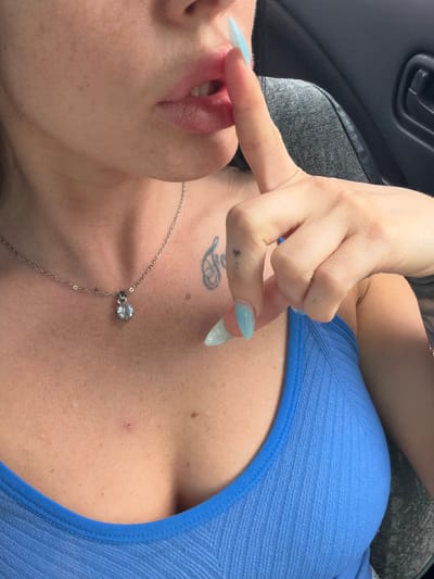 The image shows a close-up view of a person's chest and face. The person is wearing a blue, textured top. A silver necklace with a clear pendant is visible on their chest. The person has a tattoo on their upper chest, and their fingernails are long and appear to be painted a light blue color. One finger is touching their lips. The background suggests an indoor setting, possibly a car, with a dark gray handle and seating visible. There is no text visible in the image. The lighting suggests it is daytime.