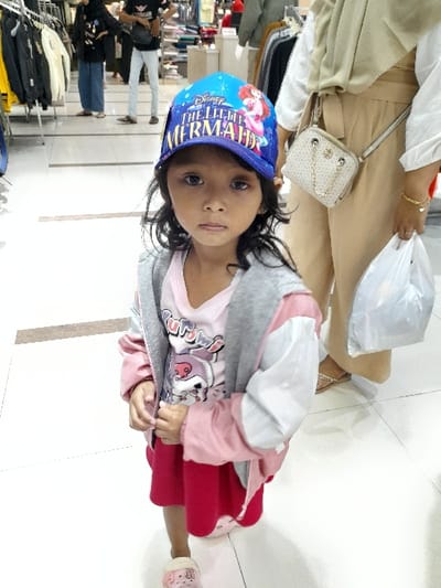 This image captures a young child positioned in the foreground of an indoor retail establishment, likely a clothing store or department store, situated within Sidoarjo, Indonesia. The precise location or store name within Sidoarjo cannot be determined from the visual information provided.
The central figure is a young individual, appearing to be 4-6 years old, with dark hair and a neutral expression, gazing directly at the camera. This child wears a blue baseball cap featuring the text "Disney THE LITTLE MERMAID" and an illustration of Ariel. The attire consists of a light pink long-sleeved shirt with a graphic, a grey zip-up hoodie, a pink and white jacket worn over the hoodie, a red skirt, and pink slippers adorned with a bow. The child's hands are clasped together in front of the body.
To the right and slightly behind the child, an adult, consistent with an adult female, is partially visible. This individual is dressed in a light beige hijab, a white long-sleeved top, and light beige trousers, carrying a light-colored patterned shoulder bag with a chain strap, and holding a white plastic shopping bag in their left hand. In the background, additional individuals are observed within the retail space: one person on the left wears a dark hijab, black top, and blue pants, while another person in the center wears a dark graphic t-shirt and light-colored pants.
The environment is characterized by bright artificial lighting and a light-colored tiled floor. Numerous clothing racks laden with various garments and displays are visible throughout the background, confirming the commercial apparel setting.