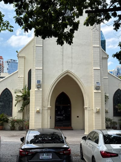 The image shows the facade of a light-colored church in Bangkok, Thailand, framed by dark green foliage from trees in the foreground. The church has a prominent arched entrance, flanked by two tall, narrow arched windows with stained glass. Above these windows, on the exterior wall, are two solar panels with attached lights. The building's architecture features decorative stone elements and pilasters. In the background, modern city buildings, including skyscrapers, are visible, indicating an urban setting. In front of the church, two cars are parked on a gravel surface. The car closer to the viewer is black and the other is white. The sky is partly cloudy.