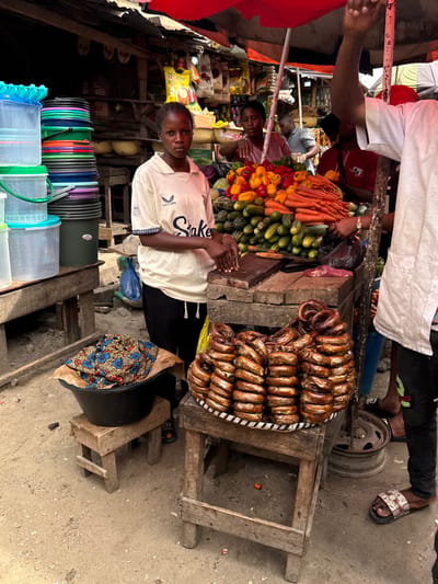 The image captures a vibrant, bustling scene within an outdoor market, likely during daytime hours. The perspective is slightly elevated, looking down into the market stalls, which are constructed from simple wooden frames.

In the center of the foreground, a young person, possibly male, stands facing the camera with a calm, neutral expression. He is wearing a light-colored, short-sleeved t-shirt with "Skake" (or "Snake") and "Castore" visible on the front. His hands are clasped together at his waist. To his left, another individual is partially visible, also looking towards the camera with hands clasped.

Behind these individuals, two main market stalls are prominently displayed. The stall on the left is piled high with a colorful array of fresh produce, including bright orange carrots, green cucumbers or zucchini, and a heap of red, yellow, and orange bell peppers. Adjacent to this vegetable stall, on the right, a large, round tray or woven mat is laden with numerous pieces of what appear to be smoked or dried fish, possibly eels, uniformly coiled into rings, presenting a dark, glossy, reddish-brown appearance.

Further back and slightly above the vegetable stall, a person dressed in a white top or robe and black trousers is partially visible, with their hands reaching into a black bag, suggesting they are handling merchandise or making a transaction. Their feet are visible, wearing sandals that appear to have some text on them. A metal wheel rim rests on the ground beneath their leg.

To the far left of the frame, a variety of household items are stacked. These include colorful plastic buckets in shades of green, blue, orange, and black, alongside clear plastic containers with colored lids. On the ground, near the dried fish stall, a large black basin or pot holds folded fabric patterned with blue, red, and white floral designs. The ground surface throughout the market area is a mix of dirt and worn concrete. In the background, behind the primary stalls, glimpses of more market activity and assorted goods can be seen, though less distinctly.