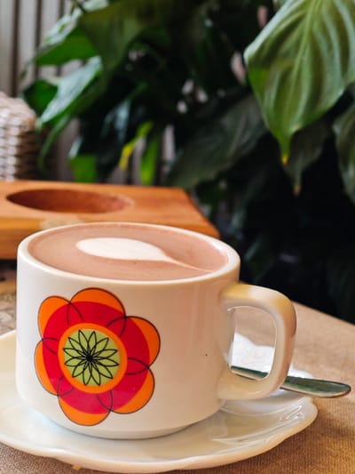 A close-up shot focuses on a white ceramic mug with a vibrant, stylized flower design. The mug is adorned with a large, multi-layered flower featuring red, orange, and green petals and a black outline. The mug contains a chocolate-colored beverage topped with a white foam art, resembling a heart shape. The mug sits on a matching white saucer, which is placed on a textured, light brown surface, possibly a tablecloth. A metallic spoon rests on the saucer, extending from beneath the mug. In the background, out of focus, are lush green foliage, suggesting an indoor plant, and a light-colored woven object, possibly a basket. Further in the background, a wooden surface with an oval indentation is visible. The overall scene evokes a cozy, cafe-like atmosphere, possibly situated in a location like Lâm Viên in Đà Lạt, Vietnam, known for its natural beauty and cafes.