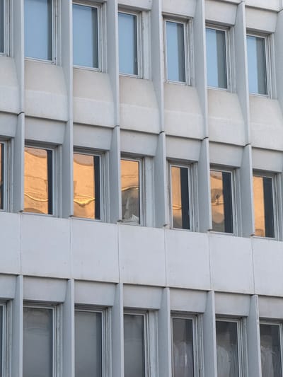 The image displays a close-up, eye-level view of the exterior of a concrete building in Tel Aviv, Israel. The facade is characterized by a repeating pattern of vertical concrete fins and recessed windows. The windows are rectangular and appear to be framed in white or light-colored material.
Some windows reflect the sky, appearing in shades of blue and gray. Other windows reflect warm, golden light, suggesting sunlight interacting with the building's interior or the surrounding environment. The texture of the concrete is rough and appears to be weathered. The overall impression is of a modern architectural style, possibly from the mid to late 20th century. There are no visible people, vehicles, or street-level elements. The focus is solely on the building's facade and its windows.
