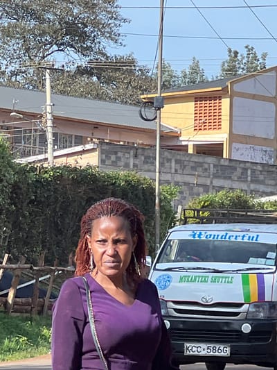 An outdoor daylight image depicts a woman in the foreground, positioned slightly to the left, with a vehicle partially visible behind her to the right. The woman has brown skin, reddish-brown braided hair styled with some small white beads, and wears a dark purple long-sleeved top with a V-neck. She carries a dark shoulder bag with a metallic chain strap. Her facial expression appears strained or disapproving, with her eyebrows furrowed and mouth slightly downturned.

Behind the woman, a white Toyota Hiace van, labeled "Wonderful" in blue script on the windshield banner and "MUKANYEKI SHUTTLE" in green lettering below, is partially visible. It has a green, purple, and yellow stripe design on its side. The vehicle bears a Kenyan license plate, "KCC 586G".

The background features a dense green hedge separating the street from a multi-story building complex. Beyond the hedge, a grey concrete block wall with a patterned design is visible. Further back, two distinct buildings are discernible. To the left, a light-colored building with a grey corrugated roof, multiple windows, and a balcony with white railings. To the right, a yellow-painted building with a dark roof and a prominent rectangular section featuring a grid pattern of orange ventilation bricks. Utility poles with wires traverse the upper portion of the scene against a clear blue sky. Trees with sparse foliage are visible in the upper background. The scene is located in the city of Nyeri, Kenya.