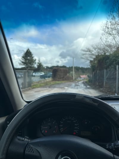 The image depicts a first-person perspective from inside a stationary vehicle in Vila Real, Portugal, looking out through the windshield. The vehicle's steering wheel, partially covered by a distressed, peeling black and grey cover, occupies the lower portion of the frame. The dashboard instrument cluster is visible, showing a tachometer on the left and a speedometer (up to 220 km/h) on the right, with several illuminated warning lights.
Beyond the windshield, the scene shows an unpaved, wet, and muddy track extending into the distance under an overcast sky with patches of blue and bright sunlight. To the left of the track, a metal gate leads to an area containing several parked vehicles, including light-colored vans, behind which a tall evergreen tree is prominent. To the right of the track, a trimmed, dark green hedge or large bush is present, followed by a metal fence and bare deciduous trees. A utility pole stands further down the track on the right. In the far background, a building with a reddish roof is faintly visible amidst more distant foliage. No people are visible.