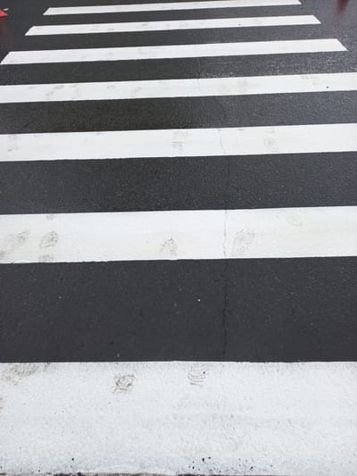 The image shows a close-up view of a pedestrian crosswalk with alternating black asphalt and white painted stripes. The crosswalk is wet, suggesting recent rain or damp conditions. Scattered across the white stripes are faint footprints, indicating that people have walked across the road. The overall lighting suggests it could be daytime, but the weather appears overcast, possibly contributing to the wetness of the road surface. The scene is a typical urban or suburban street setting. There is no visible text in the image. The context provided places this scene in Norilsk, Russia, a city known for its harsh climate and industrial environment, which could explain the wet conditions and the utilitarian nature of the crosswalk.