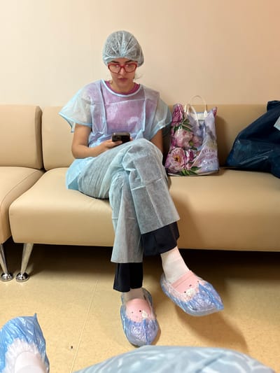 A woman is sitting on a beige couch and looking at her phone. She is wearing a blue disposable medical gown, a hairnet, red-rimmed glasses, white socks, and pink disposable shoe covers with cartoon animal designs. To her right is a shopping bag with a floral pattern and the text "TOUR DE FRANCE." To her right is a dark blue garbage bag. The scene appears to be an indoor waiting area, possibly in a medical facility. The floor is tiled in a light yellow color. There are two pairs of blue disposable shoe covers visible on the floor in front of the couch. The lighting is even and there are no obvious shadows. The overall impression is one of a sterile environment. This image was captured in Khabarovsk, Russia.