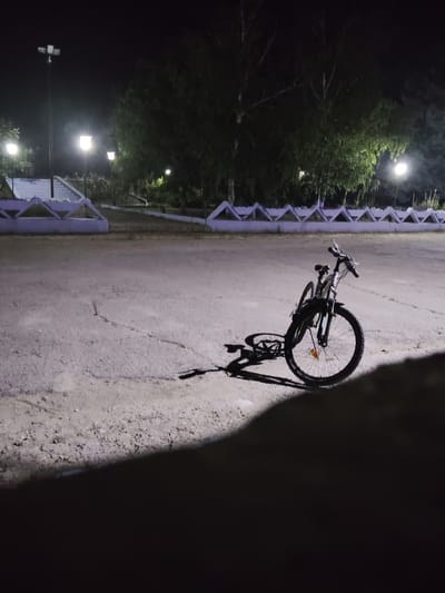 In the city of Ucrainca, Moldova, at night, a bicycle is parked on a cracked asphalt surface. The bicycle is black with silver accents and has thick tires. It is leaning against itself, with its front wheel turned to the right. A shadow of the bicycle is cast on the ground to its left. In the background, there are trees and streetlights illuminating the scene. A low, decorative fence runs along the edge of the paved area. The overall impression is one of a quiet, outdoor setting at night.