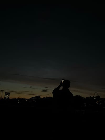 The image is a silhouetted shot of a person holding up a smartphone to take a photo. The scene appears to be at dusk or early evening. In the background, a city skyline is visible with several buildings, including a tall, modern skyscraper with lights on. The sky is a deep blue-grey, with streaks of orange and yellow clouds suggesting the last light of the sun. The overall impression is one of urban landscape at twilight. The specific location of the city of Berlin, Germany, is not discernible from the visual information present in the image.