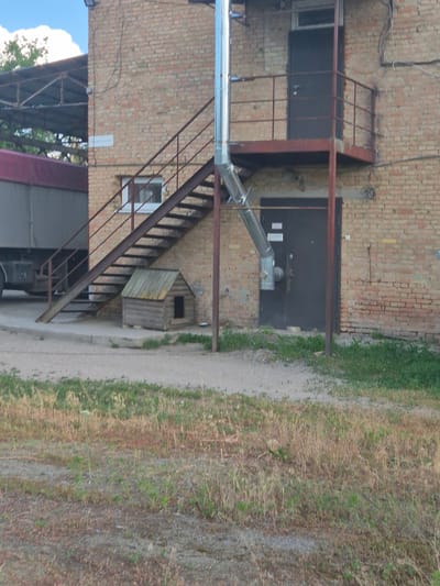 The image shows the exterior of a brick building in Sozonivka, Ukraine. A metal external staircase with a rusted railing ascends to a landing with a dark door and a balcony. A large metal pipe runs vertically along the side of the building, connected to a smaller pipe that angles downwards. Below the staircase, a small wooden doghouse is visible on a concrete surface. To the left of the building, the side of a large truck with a red tarp is visible. The ground in the foreground is a mix of gravel and dry grass. The sky is visible in the upper left corner.