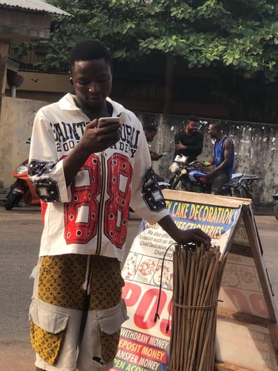 A man is looking at a cell phone and standing in front of a sign that reads "TY CAKE DECORATION." The sign also advertises withdrawal, deposit, and transfer money. The man is wearing a white shirt with a large red "88" on the front and gold and brown patterned pants. In the background, there are other people and motorbikes. Green foliage is visible on the left. The scene is outdoors, and the lighting suggests it is daytime.