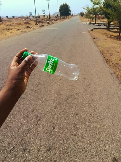 The image shows a hand holding an empty Sprite bottle on a paved road. The location is in Rafin Pa, Nigeria, as evidenced by the typical vegetation and landscape. The road stretches into the distance, lined with trees and dry, grassy areas. There is some development visible in the background.