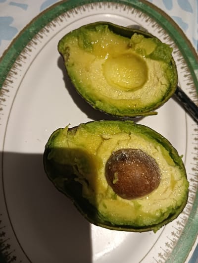 The image shows two halves of a ripe avocado, cut lengthwise, resting on a white oval plate with a decorative green and gold rim. The avocado flesh is a vibrant green, with a creamy texture. One half contains a large, brown pit, dusted with what appears to be sugar crystals. The other half is pitted and has a slightly concave surface. Small water droplets glisten on the avocado flesh, suggesting it is fresh or has been recently washed. The lighting indicates it might be daytime, possibly indoors, as evidenced by the shadow cast on the plate by one of the avocado halves. A dark object, possibly a utensil, is partially visible to the right of the upper avocado half. The plate is set on a surface with a patterned fabric, possibly a tablecloth, featuring blue and white elements. The overall scene is a simple, appetizing presentation of the fruit, prepared for consumption. The location context is Dubai, United Arab Emirates.