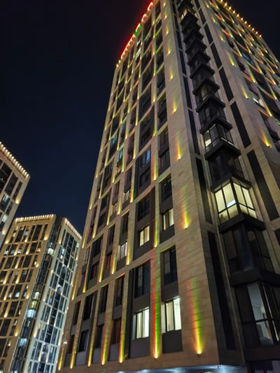 The image depicts a nighttime view of modern high-rise apartment buildings in Almaty, Kazakhstan. The buildings are illuminated with colorful vertical lights in red, green, and yellow, casting a vibrant glow against the dark sky. The architecture features a contemporary design with many windows and balconies. In the distance, another similar building is partially visible. The sky is clear and dark, with a few faint stars visible. There are no people or vehicles clearly visible in the frame. The scene conveys a sense of urban nocturnal beauty and architectural illumination.