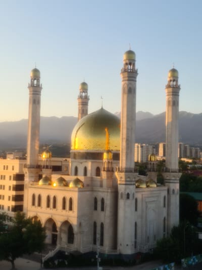 This image shows a view of the Central State Mosque in Almaty, Kazakhstan, bathed in the soft light of either sunrise or sunset. The mosque is a prominent white structure with a large, gleaming golden dome at its center, surrounded by four smaller golden domes. Four tall minarets, also capped with golden domes, flank the main building.

The mosque is situated within a city environment, with modern high-rise buildings visible in the background, suggesting an urban setting. The mountains of the Tian Shan range loom in the distance, partially obscured by haze. In the foreground, a large green tree stands to the left of the mosque, and part of a street or plaza is visible with some low-lying greenery and architectural elements. The sky is a clear, pale blue, indicating fair weather. No people or animals are visible in the frame. The lighting suggests a time of day when artificial lights would not yet be illuminated, reinforcing the impression of early morning or late evening.